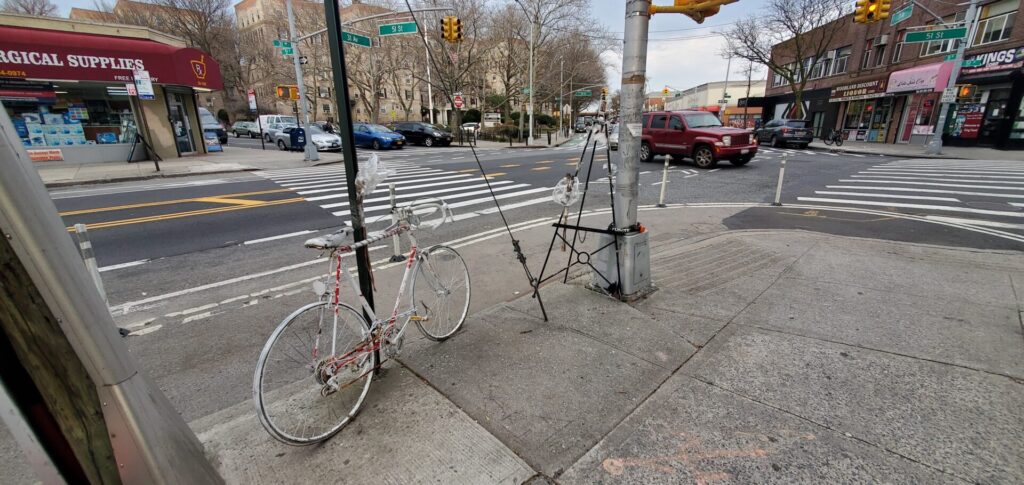 White ghost bike next to empty easel attached to street signs on 31 Avenue at 51 Street in Woodside, Queens.
