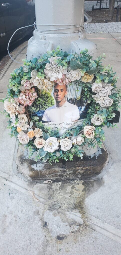 Floral wreath with a young man's picture in the middle of it and a sign that reads "In Memory of Vickram Sewsanka 1994 - September 3, 2020"